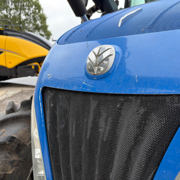 Blue tractor front grille with emblem, close-up shot; a yellow agricultural machine is visible in the background in an outdoor lot.