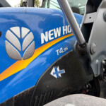 Close-up of a blue New Holland tractor body with white lettering and an orange stripe, hydraulic arm in the foreground.