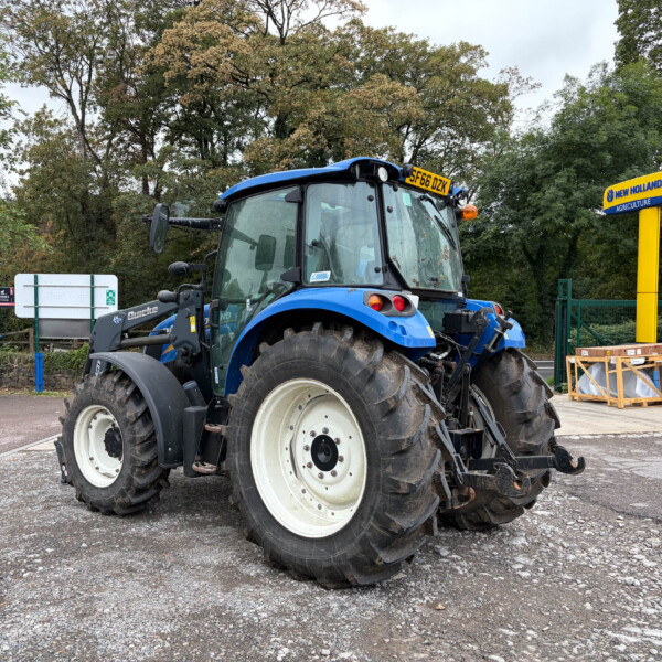 Blue New Holland tractor on a gravel lot with large tires and a yellow machinery sign in the background.] ,