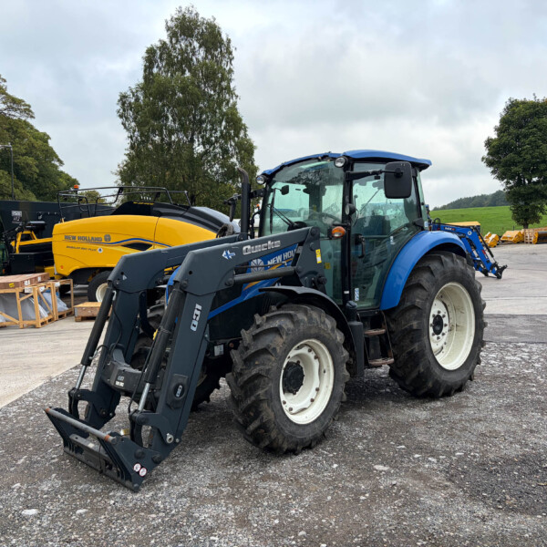 Blue tractor with a front loader in a construction yard, surrounded by other equipment and a yellow machine nearby.