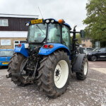 Front view of a blue tractor with oversized muddy tires parked in a parking lot near a building with a sign visible in the background, license plate SF66 DZK on the cab roof.