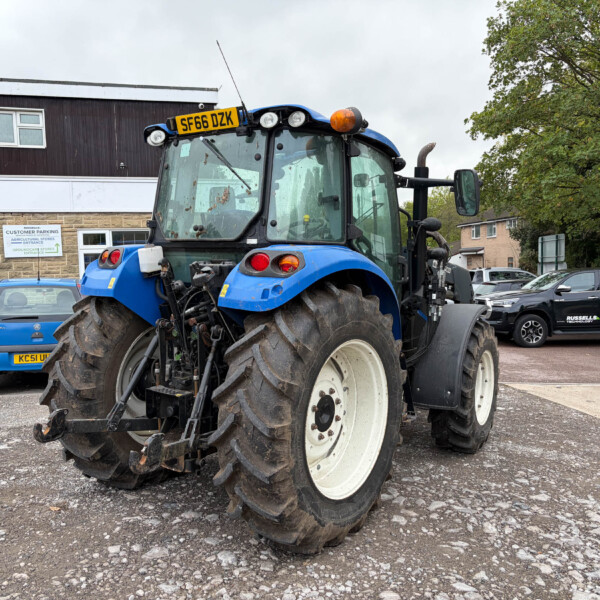 Front view of a blue tractor with oversized muddy tires parked in a parking lot near a building with a sign visible in the background, license plate SF66 DZK on the cab roof.