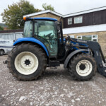 Blue tractor with front loader parked in a gravel lot beside a brick-and-white building; two white delivery vans in the background.
