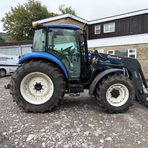 Blue tractor with front loader parked in a gravel lot beside a brick-and-white building; two white delivery vans in the background.