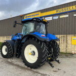 Blue New Holland tractor with large white-walled tires parked outside a dark gray industrial building with a yellow and blue Russell sign above it.