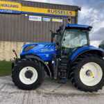 Blue New Holland tractor parked in front of a dark brown building with a bright yellow-and-blue 'NEW HOLLAND AGRICULTURE' sign visible above.