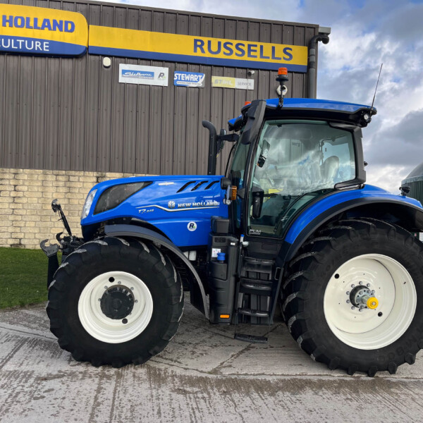 Blue New Holland tractor parked in front of a dark brown building with a bright yellow-and-blue 'NEW HOLLAND AGRICULTURE' sign visible above.