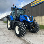 Blue New Holland agricultural tractor parked outside a storefront with a yellow sign reading 'New Holland Agriculture' on a gray corrugated wall.