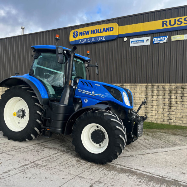 Blue New Holland tractor in front of a gray corrugated metal building with a yellow-and-blue New Holland Agriculture sign.