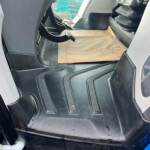 Driver's side footwell in a vehicle showing pedals and a brown paper floor mat; blue coolant reservoir cap visible nearby