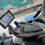 Inside a heavy machinery cab with a touchscreen tablet, joystick controls, and a construction site outside with green tarps and concrete blocksyje (image shows interior and work area)
