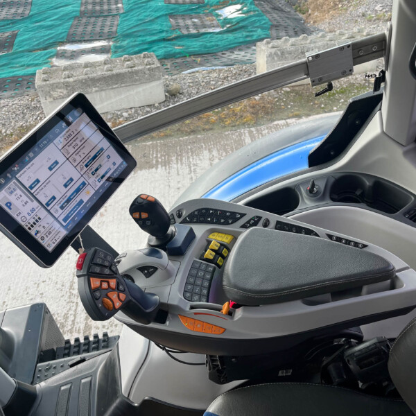 Inside a heavy machinery cab with a touchscreen tablet, joystick controls, and a construction site outside with green tarps and concrete blocksyje (image shows interior and work area)