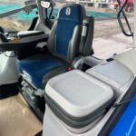 Inside a modern tractor/bulldozer cabin with a blue-upholstered driver seat, steering wheel, pedals, and a touchscreen display.