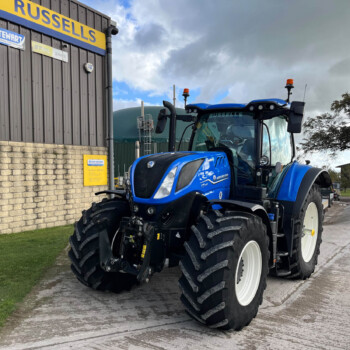Blue New Holland tractor parked outdoors near a gray industrial building with a large yellow Russell sign and smaller company logos on the wall