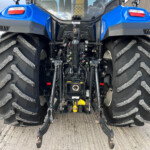 Rear view of a blue agricultural tractor with oversized tread tires, exposed hydraulic controls and three-point hitch on a concrete surface.