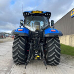 Front view of a blue New Holland tractor with oversized tires at a farm equipment yard, building in background with New Holland sign.