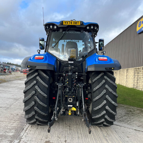 Front view of a blue New Holland tractor with oversized tires at a farm equipment yard, building in background with New Holland sign.