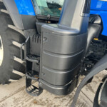 Close-up of a blue tractor showing the black stepping platform and a large treaded front wheel with a white rim and blue body panel nearby.
