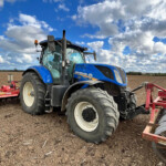 Blue New Holland tractor parked in a tilled field under a bright sky with scattered clouds. Alternative 1