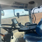 Interior view of a tractor cabin showing the steering wheel, blue cushioned seat, and dashboard controls near the windshield.