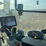 Inside view of a tractor cab showing a joystick, a touchscreen display, and the instrument panel with numerous colored buttons; plowed field visible through the windshield.