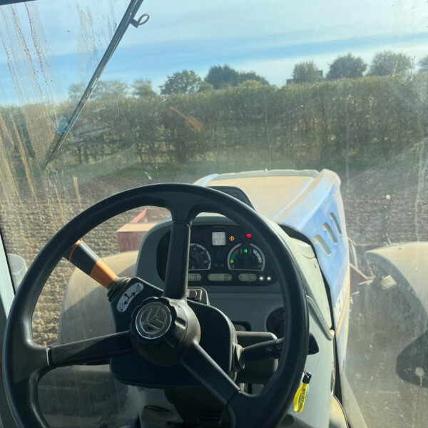 View from the tractor cab: steering wheel in foreground, dashboard gauges, and a dirt farm field visible through a dusty windshield.