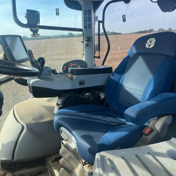 Interior of a blue tractor cab showing the driver's seat, steering wheel, and control panel with buttons and a screen; fields seen through the window.