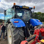 Blue agricultural tractor with orange beacon lights, license plate YX21 XLK, and red implements attached in a dirt field under a cloudy sky, with a person beside the cab door for scale.
