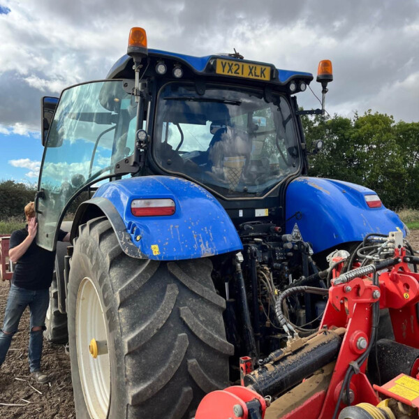 Blue agricultural tractor with orange beacon lights, license plate YX21 XLK, and red implements attached in a dirt field under a cloudy sky, with a person beside the cab door for scale.