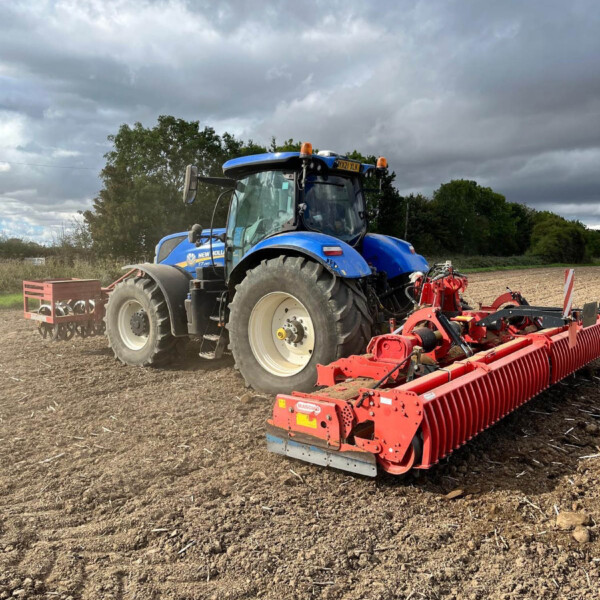 Blue tractor with a red farming implement attached, on a plowed field under a cloudy sky.