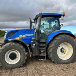Blue New Holland tractor with large tires parked on a plowed field under a cloudy sky, showing the cab and orange beacon.