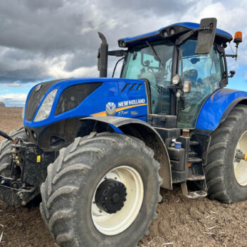 Blue New Holland T7 tractor parked on tillable soil under cloudy skies, front view emphasizing large tires and cab.