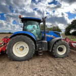 Blue New Holland tractor parked on a tilled field under a cloudy sky, ready for farming chores.