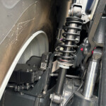 Close-up of a car's suspension with a coil spring and shock absorber beside a wheel tube area near the tire edge.
