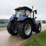 Blue agricultural tractor with oversized rear tires parked on a wet concrete lot, overcast sky and distant trucks in the background.
