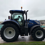 Blue New Holland tractor with large front tires and orange beacon on an overcast day, seen from the front-left side.