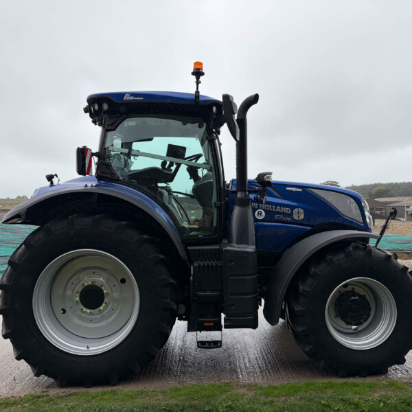 Blue New Holland tractor with large front tires and orange beacon on an overcast day, seen from the front-left side.