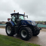Blue New Holland tractor on a damp construction site under a cloudy sky, with grassy area in the foreground and distant buildings on the horizon.