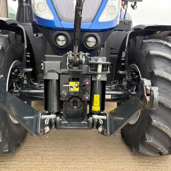 Close-up of a New Holland tractor front hitch with hydraulic linkage and dual large tread tires on either side.