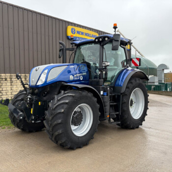 Blue New Holland tractor with large off-road tires parked near a gray industrial building and brick wall, beacon light on top, and missing front attachment on the ground nearby.