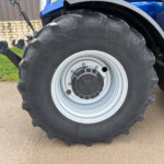 Close-up of a tractor rear tire and wheel with hitch, outdoor setting against a brick wall and grass in the background.