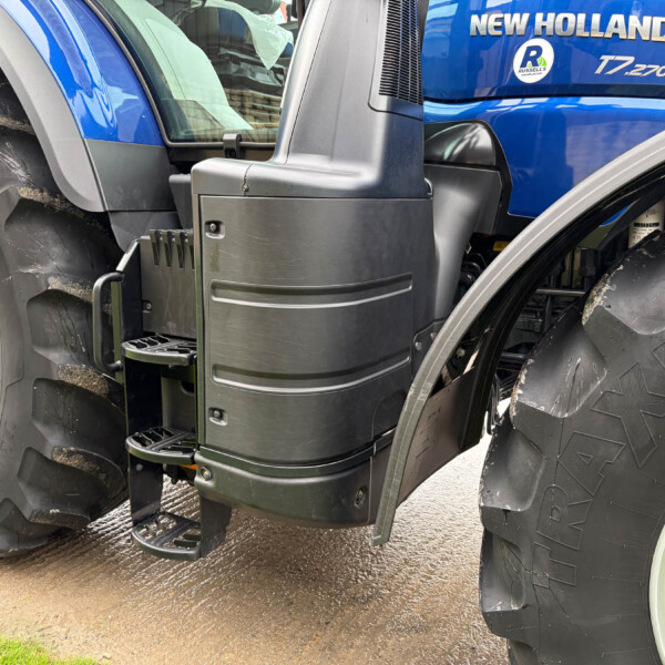 Close-up of a blue New Holland tractor showing the entry steps, black side panel, and a large muddy tire with the cab and window reflections visible.