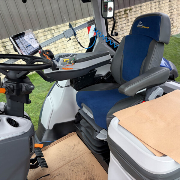 Interior of a modern tractor cabin with a blue-gray seat, steering wheel, mounted tablet, and control panel; a cardboard box sits in the foreground