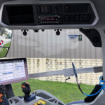 Interior of a truck cab with a mounted touchscreen display showing vehicle data and controls above the dashboard path to a driver joystick and windshield view behind.