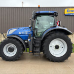 Blue New Holland tractor shown from the front-left, featuring large tires, in front of a brown wooden wall with a New Holland Agriculture sign to the right.