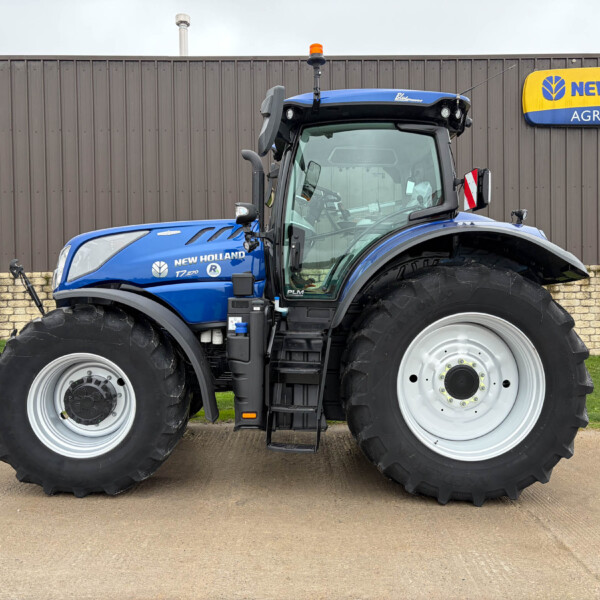 Blue New Holland tractor shown from the front-left, featuring large tires, in front of a brown wooden wall with a New Holland Agriculture sign to the right.