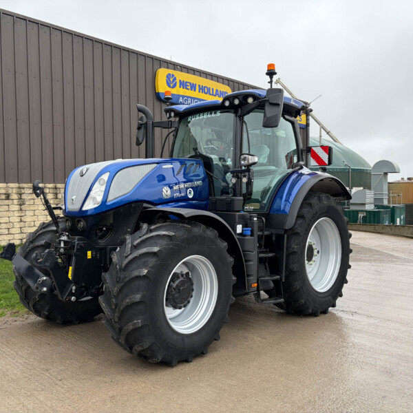 Blue New Holland tractor parked beside a brown metal building with a yellow 'New Holland Agriculture' sign above it socalled in view of the photo.