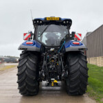 Front view of a large blue tractor in an outdoor yard, facing the camera, with a corrugated metal building on the right.