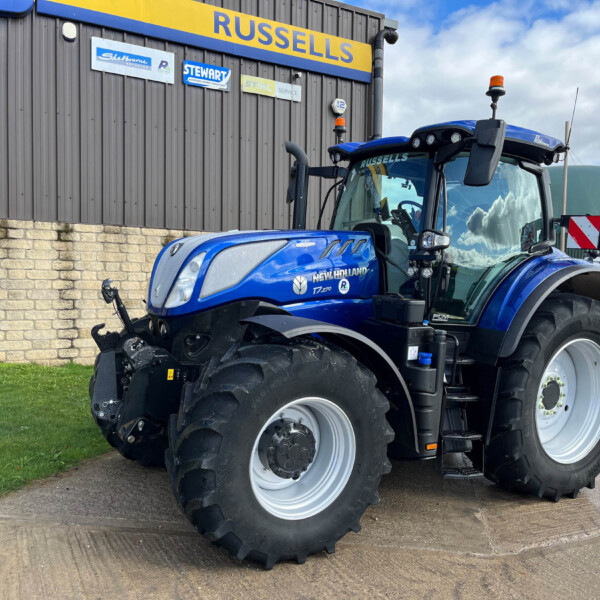Blue New Holland tractor parked outside a grey building with 'RUSSELLS' signage, large wheels and chrome exhaust visible.