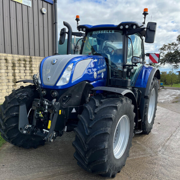 Blue agricultural tractor with oversized front and rear tires parked beside a brick wall and industrial building, orange warning lights visible on top.